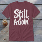 Maroon short-sleeved t-shirt with white text reading "Still A-Goin'" laid out on a weathered wooden surface next to a glass of iced tea.