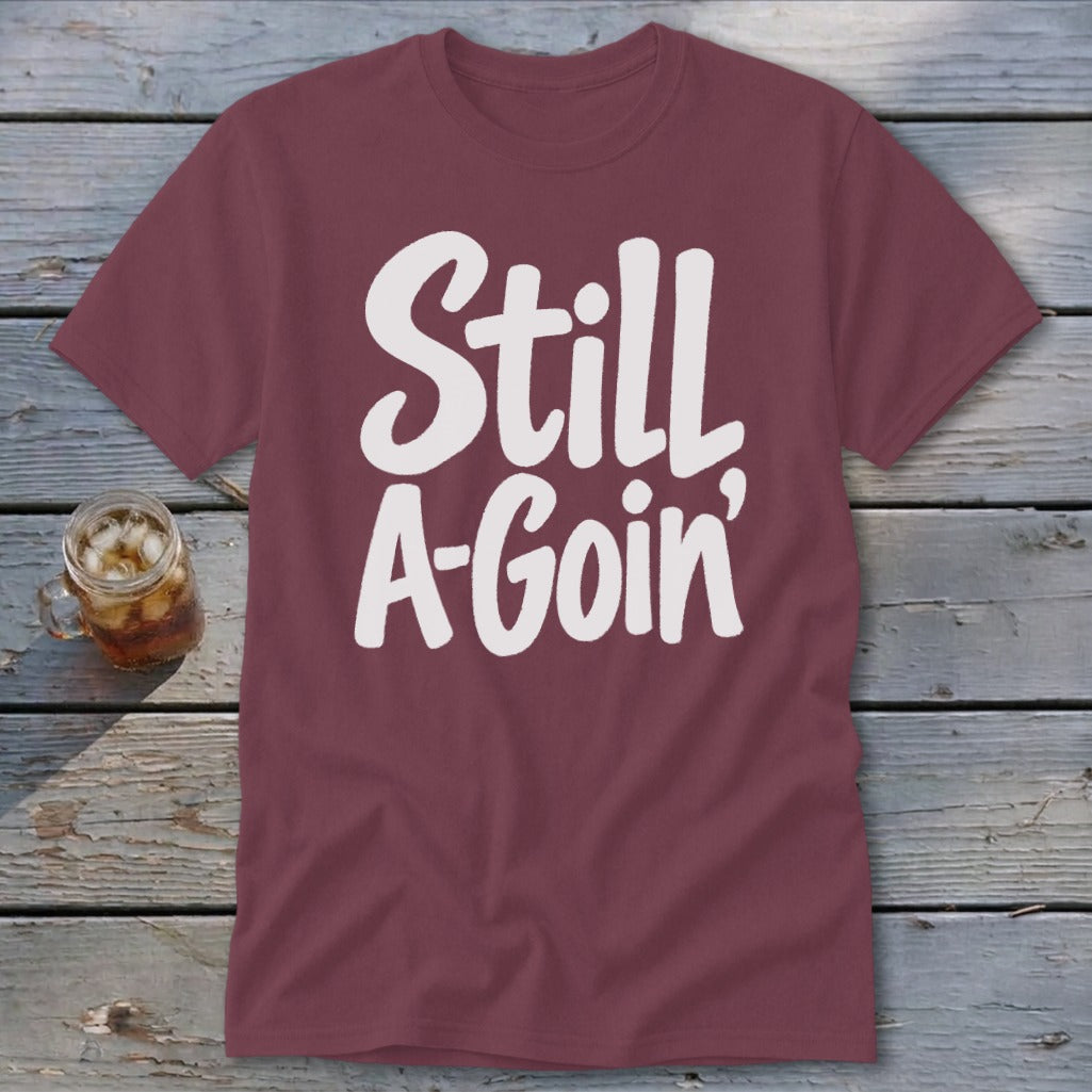 Maroon short-sleeved t-shirt with white text reading "Still A-Goin'" laid out on a weathered wooden surface next to a glass of iced tea.