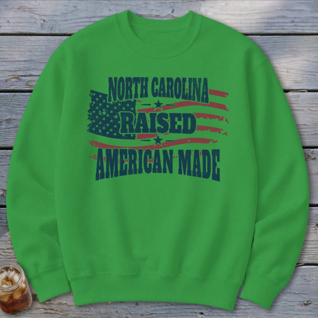 A bright green crewneck sweatshirt featuring a distressed American flag graphic with text reading North Carolina Raised American Made.