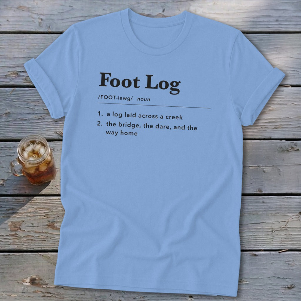 Light blue t-shirt featuring dictionary-style text defining "Foot Log" laid on a wooden surface.