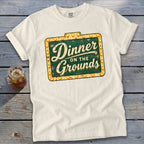 Cream short-sleeved t-shirt with a vintage sign graphic reading Dinner on the Grounds, lying on wood.