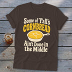 Brown t-shirt featuring a graphic of yellow cornbread in a skillet and text reading Some of Y'all's Cornbread Ain't Done in the Middle.