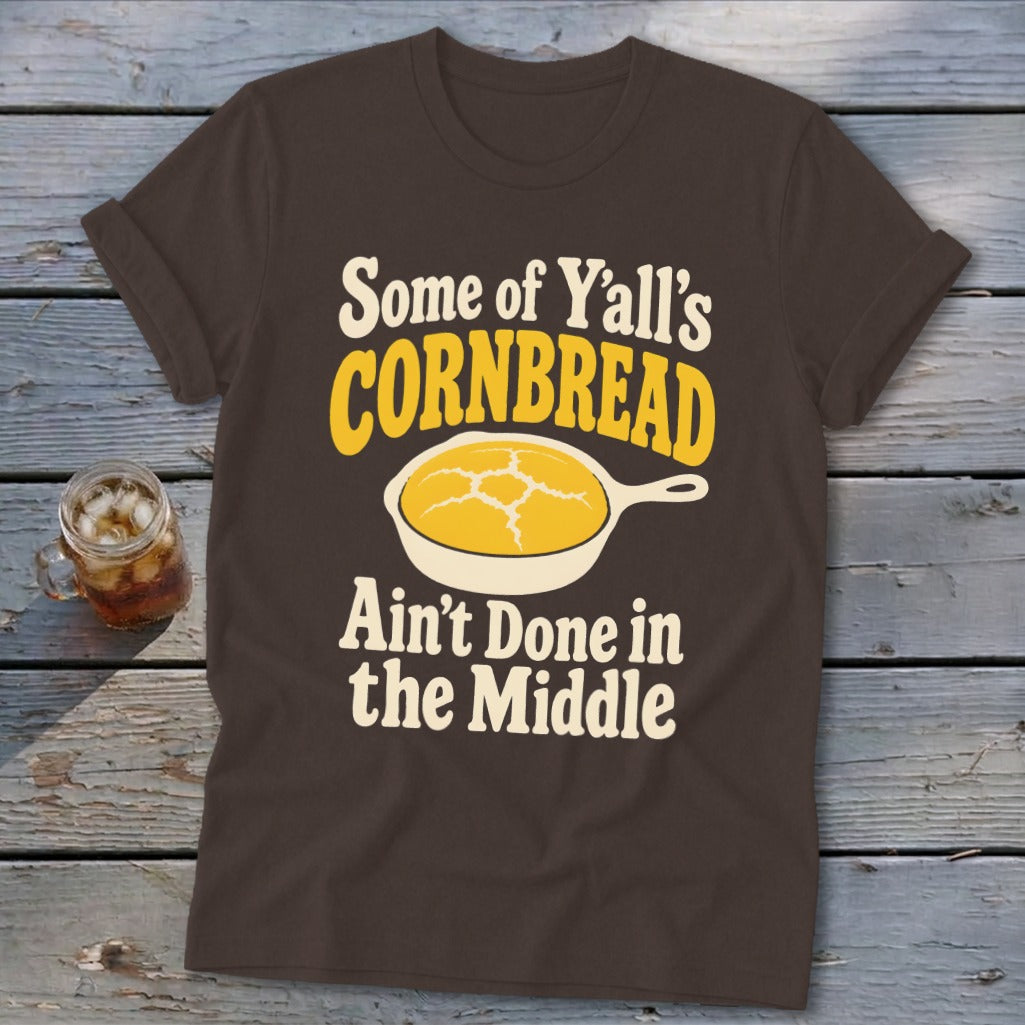 Brown t-shirt featuring a graphic of yellow cornbread in a skillet and text reading Some of Y'all's Cornbread Ain't Done in the Middle.