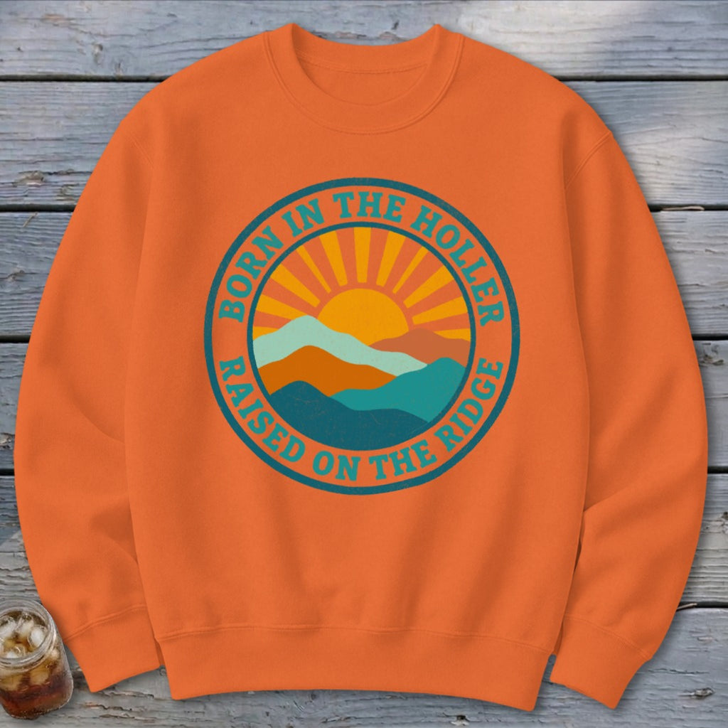 An orange crewneck sweatshirt featuring a circular graphic with mountains and the text "BORN IN THE HOLLER RAISED ON THE RIDGE".