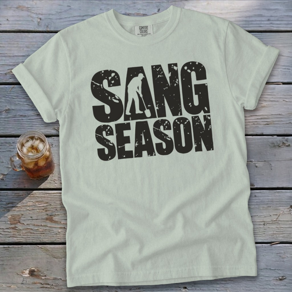 Light green t-shirt with distressed black text reading SANG SEASON laid on a rustic wooden table.
