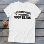 White t-shirt with black text reading THIS THANKSGIVING IS SPONSORED BY SOUP BEANS laid on a wooden background.