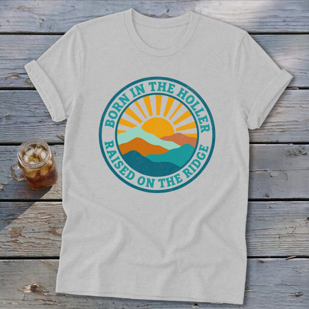 A grey t-shirt featuring a circular graphic with mountains, a rising sun, and the text BORN IN THE HOLLER RAISED ON THE RIDGE.