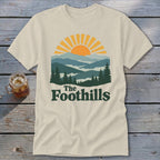 Cream t-shirt with "The Foothills" text and a sunrise over layered mountains.