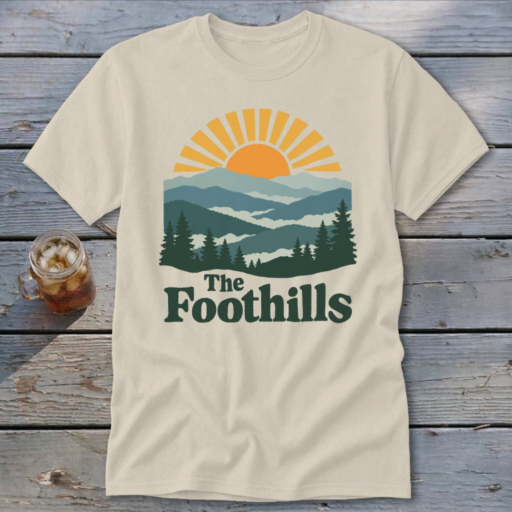 Cream t-shirt with "The Foothills" text and a sunrise over layered mountains.