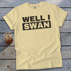 A light yellow t-shirt with black text reading "WELL I SWAN" is laid flat on a gray wooden background next to a glass of iced tea.