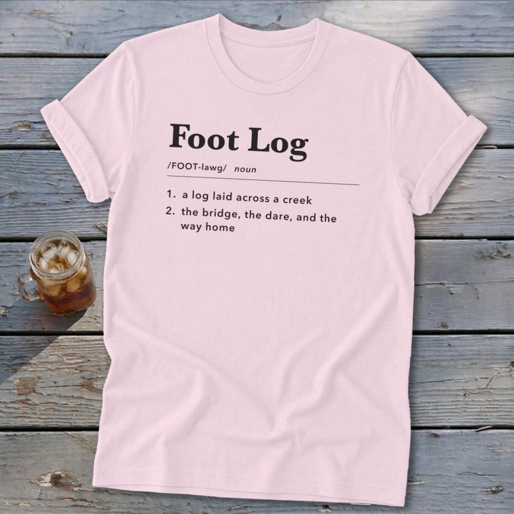 A light pink t-shirt with black text defining the term "Foot Log" laid flat on a wooden surface.