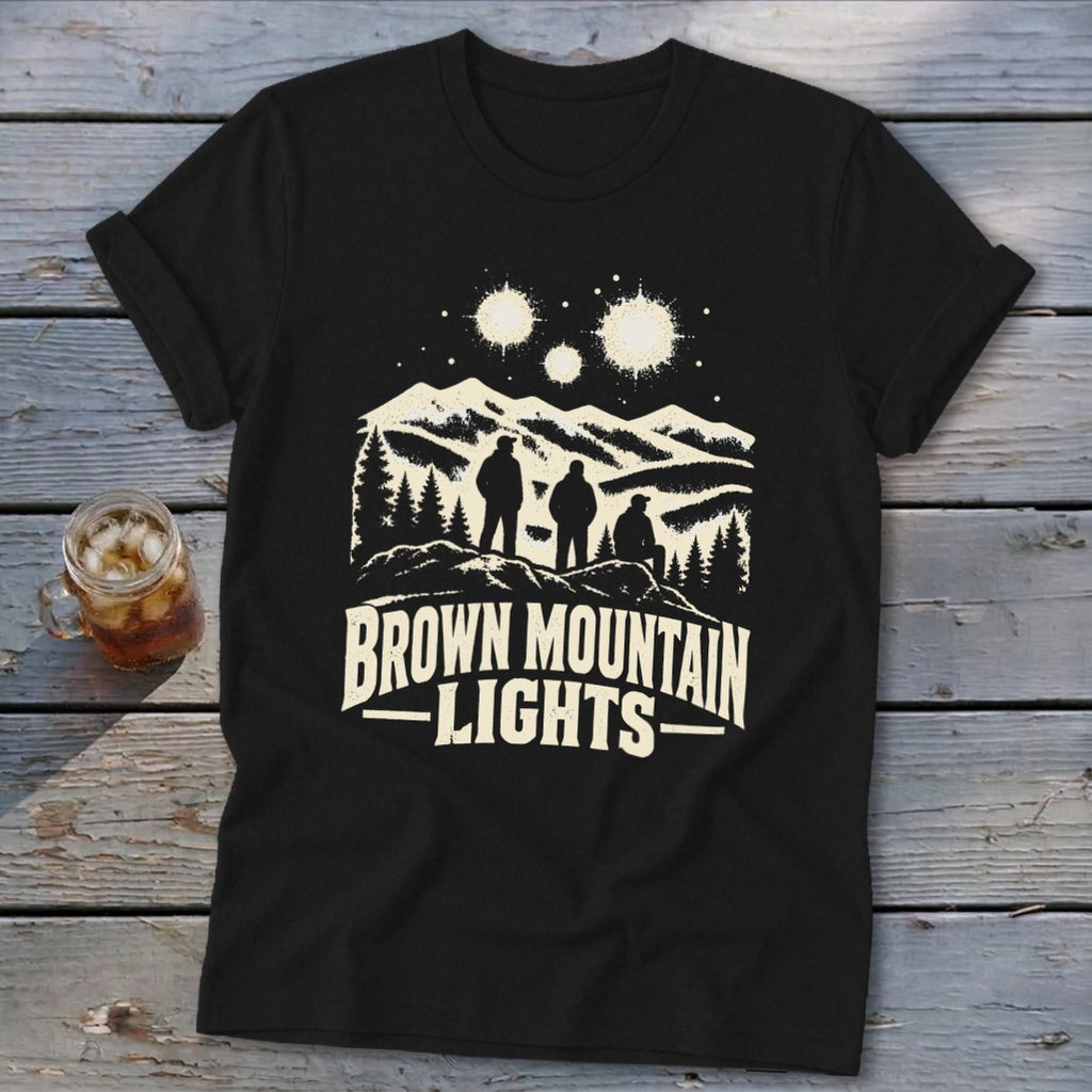 A black short-sleeved t-shirt featuring a graphic of three figures viewing glowing orbs over mountain peaks with the text Brown Mountain Lights.