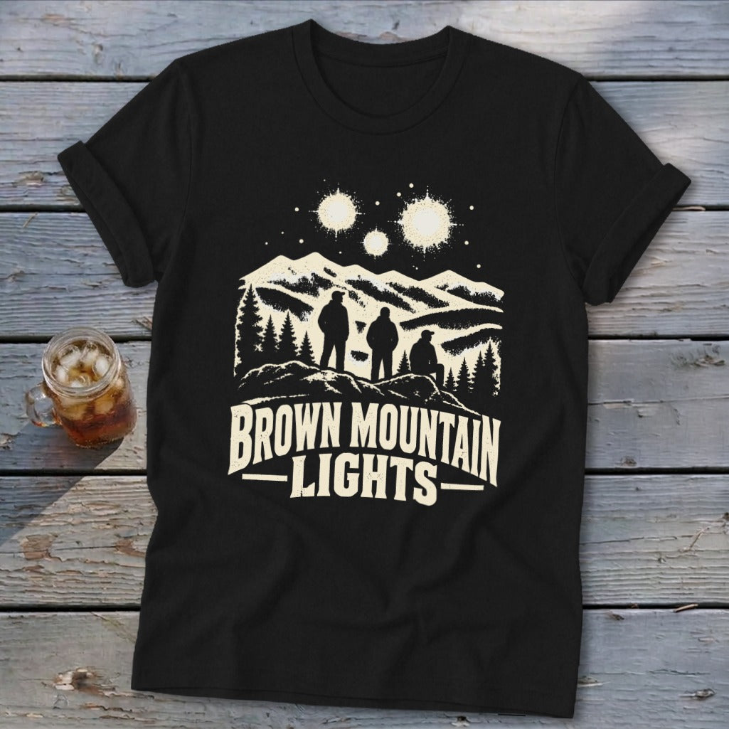 A black short-sleeved t-shirt featuring a graphic of three figures viewing glowing orbs over mountain peaks with the text Brown Mountain Lights.