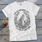 A white t-shirt featuring a black graphic of a bird perched on a fence, surrounded by corn stalks and ferns, with the text WHEN THE WHIPPOORWILL CALLS IT'S TIME TO PLANT CORN.