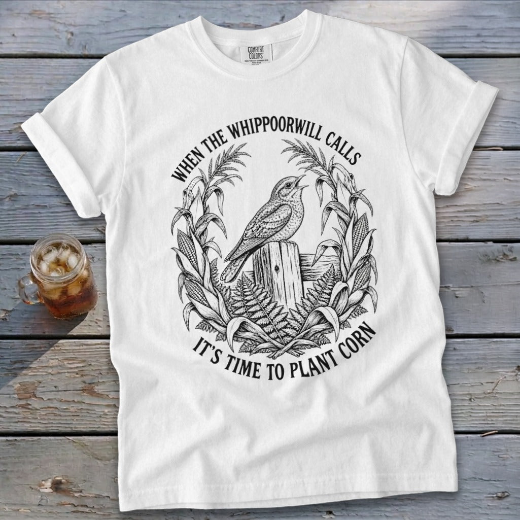 A white t-shirt featuring a black graphic of a bird perched on a fence, surrounded by corn stalks and ferns, with the text WHEN THE WHIPPOORWILL CALLS IT'S TIME TO PLANT CORN.