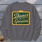 A dark gray crewneck sweatshirt featuring a distressed vintage-style green sign that reads Dinner on the Grounds.
