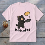 A light pink short-sleeve t-shirt features a cartoon black bear flipping a pancake from a frying pan above the word hoecakes.