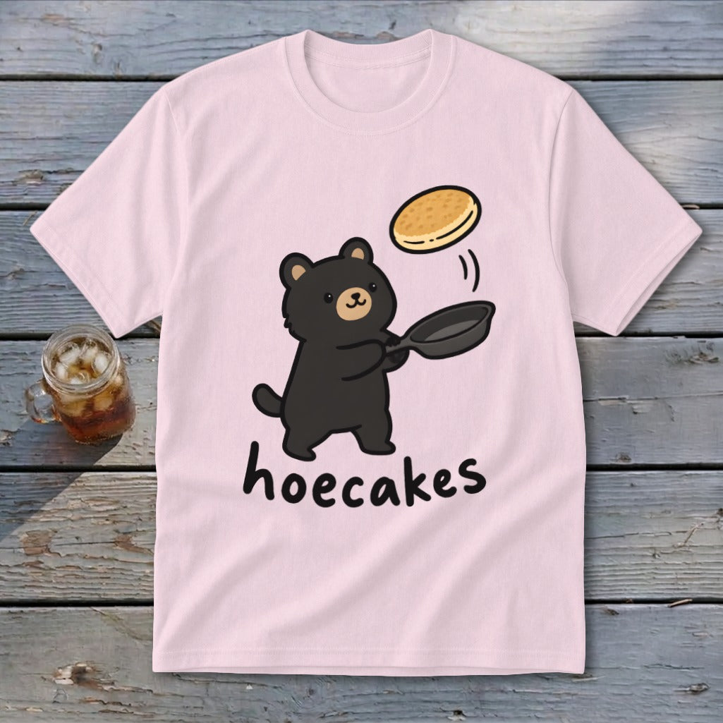 A light pink short-sleeve t-shirt features a cartoon black bear flipping a pancake from a frying pan above the word hoecakes.