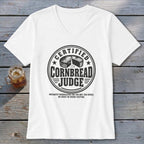 A white v-neck t-shirt displays a black graphic featuring text about certified cornbread judges on a wooden background.
