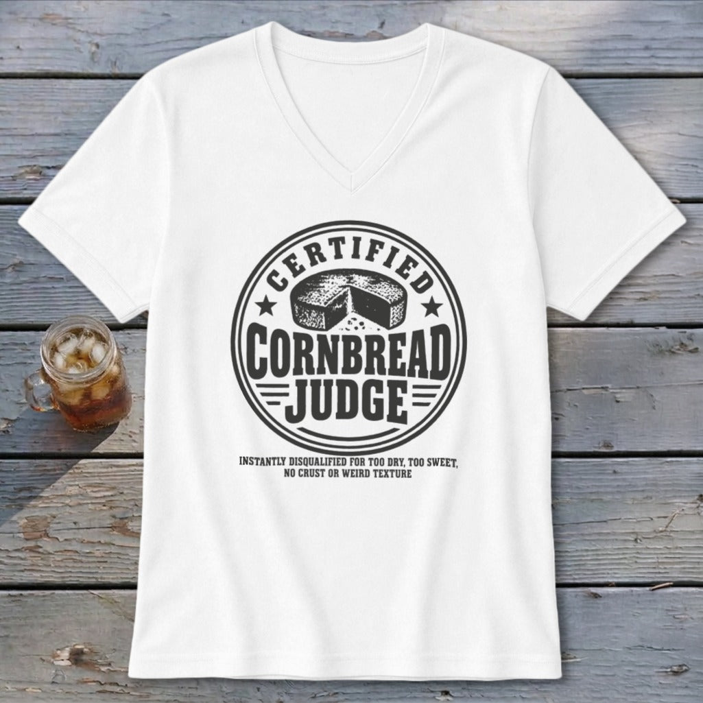 A white v-neck t-shirt displays a black graphic featuring text about certified cornbread judges on a wooden background.