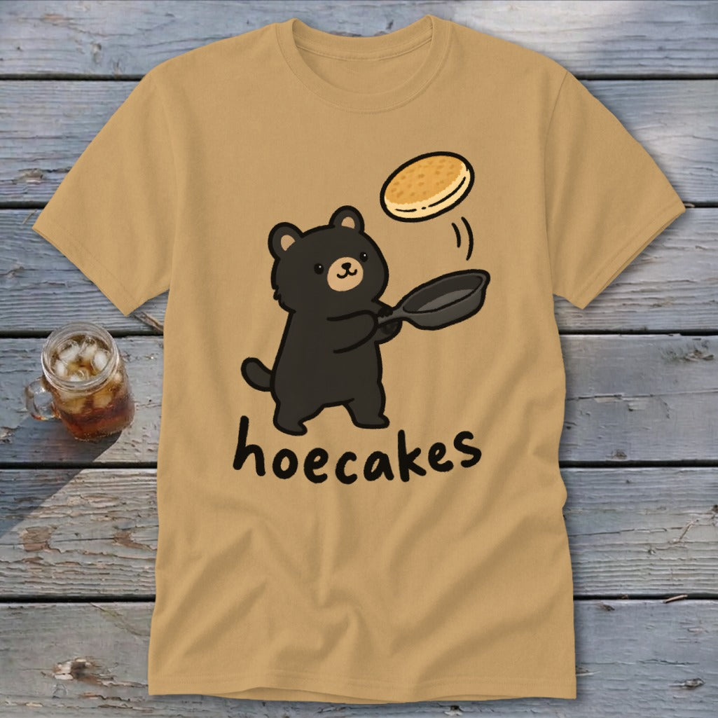 A tan short-sleeve t-shirt features a cartoon black bear flipping a pancake in a pan, with the text hoecakes underneath.