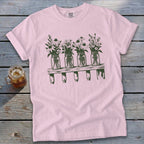 A light pink t-shirt with a green graphic of four mason jars filled with wildflowers resting on a wooden shelf, laid flat on weathered wood next to a glass of iced tea.