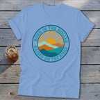 Light blue t-shirt featuring a retro mountain and sun graphic with the text Born in the Holler Raised on the Ridge.