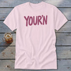 Light pink t-shirt with "YOUR'N" in purple lettering, next to a glass of iced tea.