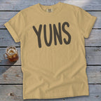 Tan t-shirt with "YUNS" text in dark gray, next to a glass of iced tea.
