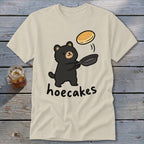 Beige t-shirt featuring a cute cartoon bear flipping a pancake above a frying pan with the text hoecakes.
