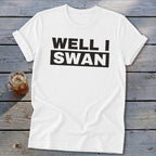 A white t-shirt with rolled sleeves lies on a rustic wooden surface next to an iced drink, featuring the black text WELL I SWAN printed across the chest.