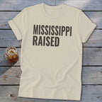 Light beige t-shirt with "MISSISSIPPI RAISED" text on a wooden surface.