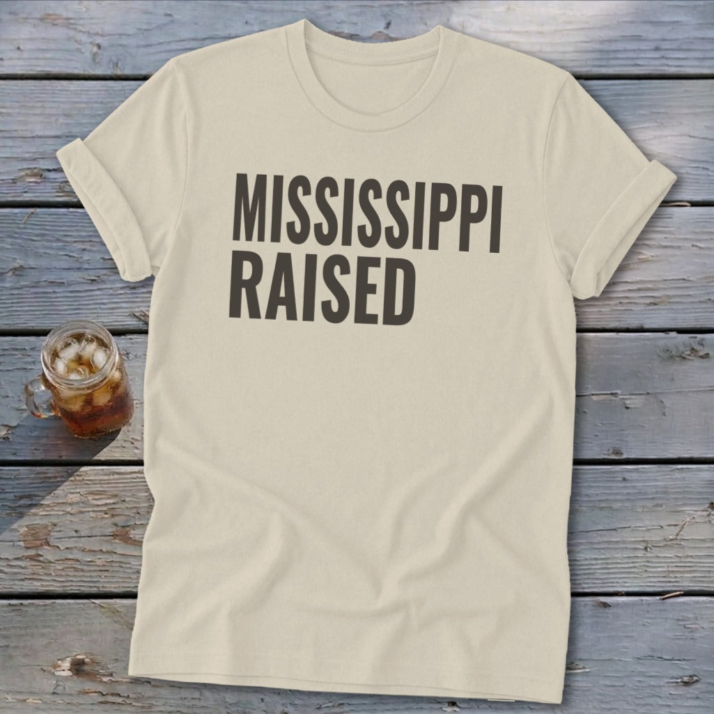 Light beige t-shirt with "MISSISSIPPI RAISED" text on a wooden surface.