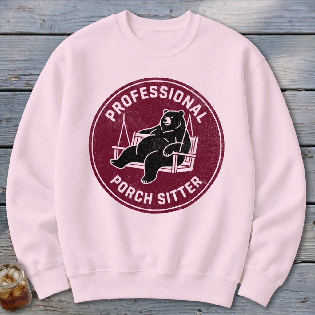 Light pink crewneck sweatshirt featuring a maroon circular graphic of a bear relaxing on a porch swing, with the text Professional Porch Sitter.