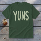 Dark green t-shirt with "YUNS" in light green font on a wooden surface.