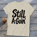 Beige crewneck t-shirt with rolled sleeves featuring black cursive text reading Still A-Goin'.