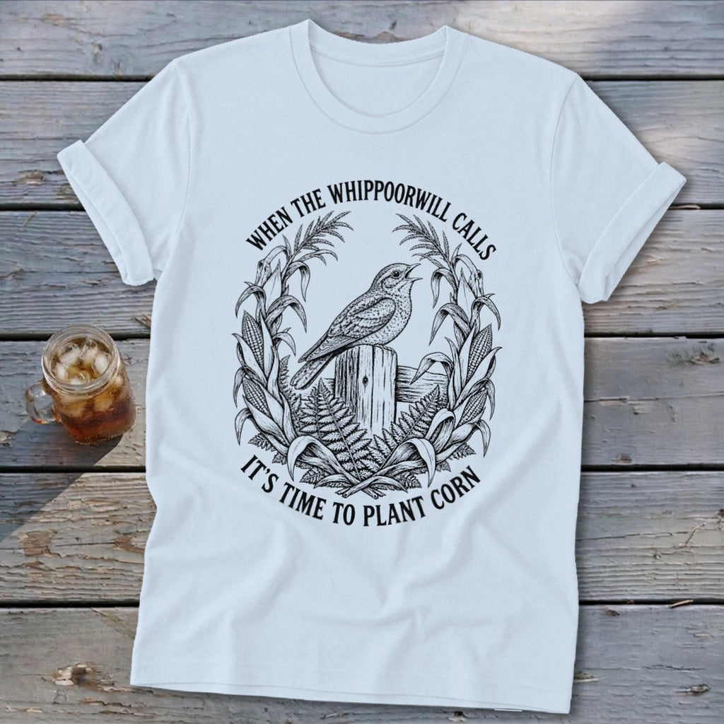 A white crewneck t-shirt with a vintage graphic of a bird on a fence surrounded by corn stalks and text that reads when the whippoorwill calls it is time to plant corn.