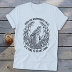 A white crewneck t-shirt with a vintage graphic of a bird on a fence surrounded by corn stalks and text that reads when the whippoorwill calls it is time to plant corn.
