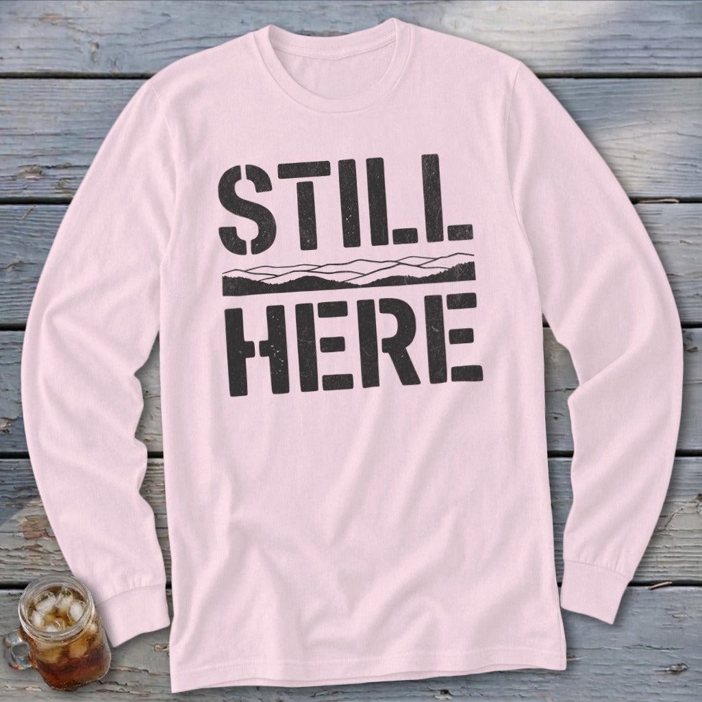 Light pink long sleeve shirt with black text "STILL HERE" and a mountain outline.
