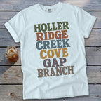 A light blue t-shirt with rolled sleeves lies flat on a weathered wooden surface, featuring colorful stacked text that reads HOLLER RIDGE CREEK COVE GAP BRANCH.