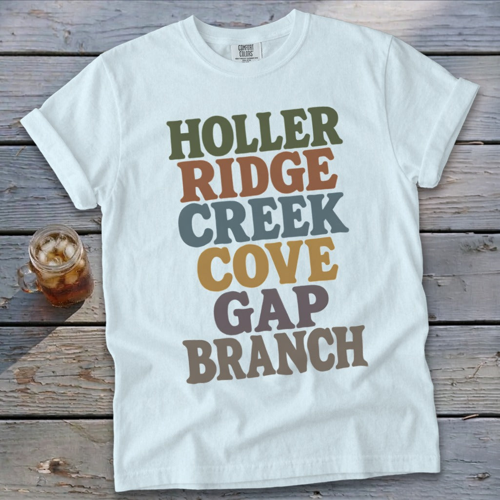 A light blue t-shirt with rolled sleeves lies flat on a weathered wooden surface, featuring colorful stacked text that reads HOLLER RIDGE CREEK COVE GAP BRANCH.