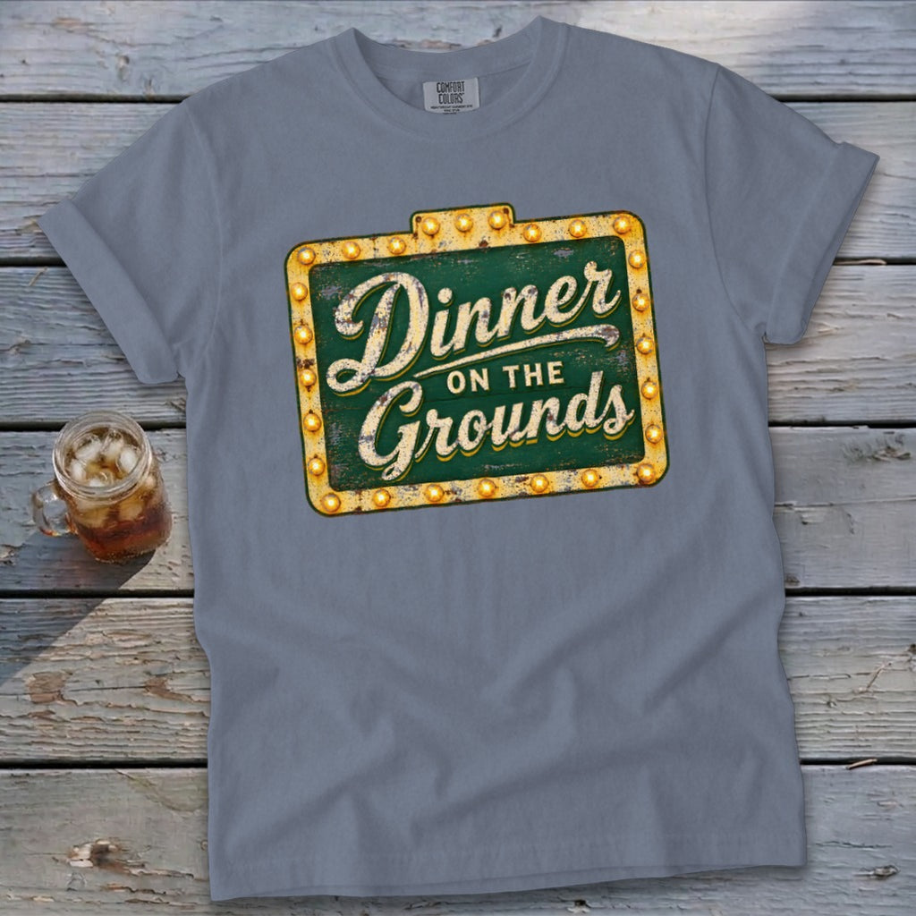 Blue-gray crewneck t-shirt with a vintage sign graphic reading "Dinner on the Grounds" laid flat on a rustic wooden surface.