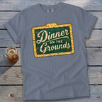 Blue-gray crewneck t-shirt with a vintage sign graphic reading "Dinner on the Grounds" laid flat on a rustic wooden surface.