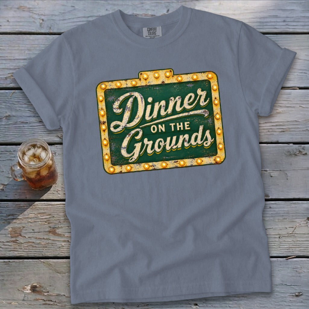 Blue-gray crewneck t-shirt with a vintage sign graphic reading "Dinner on the Grounds" laid flat on a rustic wooden surface.
