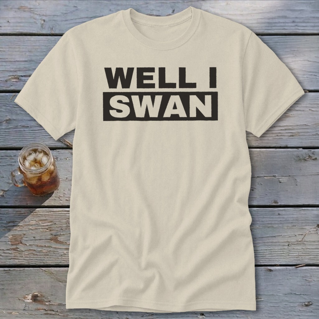 A cream-colored t-shirt with the black text Well I Swan centered on the chest, laid on a wooden surface next to an iced drink.