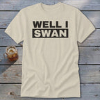 A cream-colored t-shirt with the black text Well I Swan centered on the chest, laid on a wooden surface next to an iced drink.