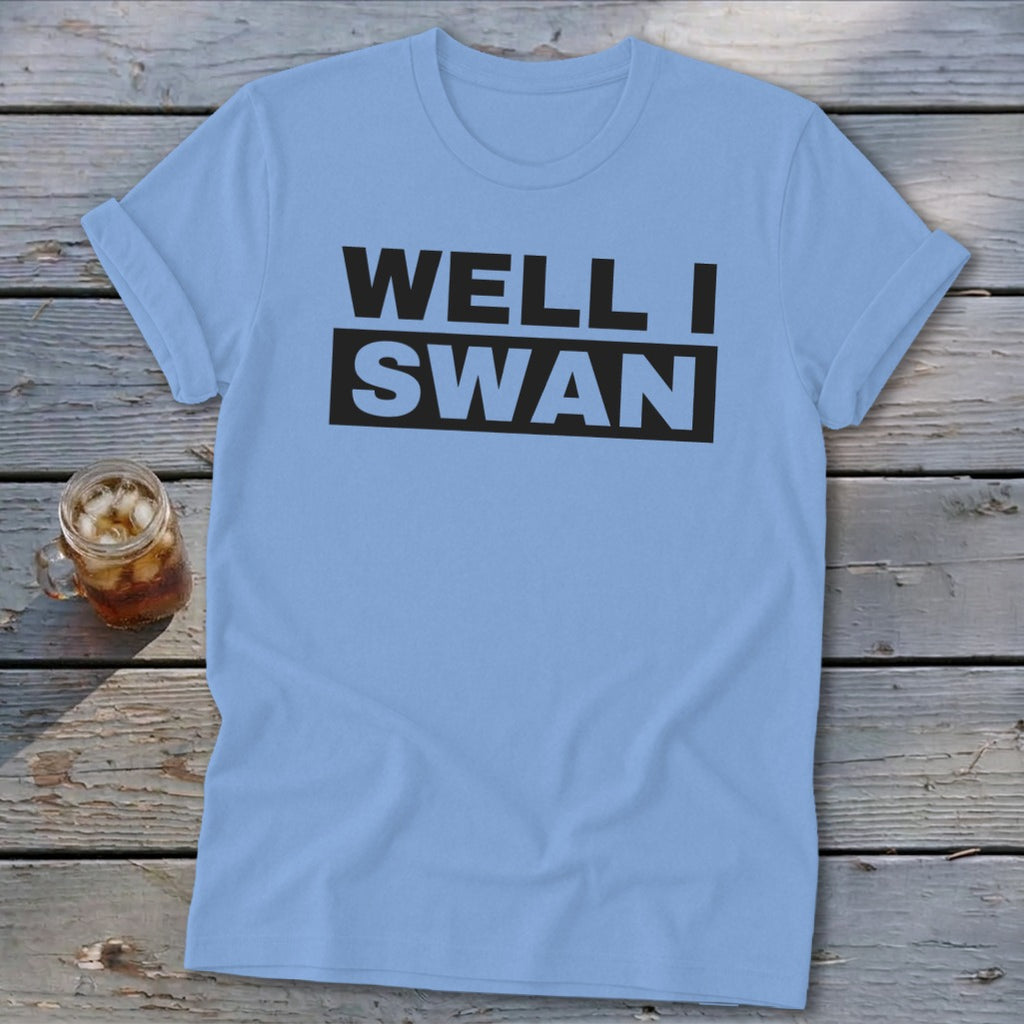 Light blue short-sleeve t-shirt with the text "WELL I SWAN" printed in black on the chest, lying on a wooden surface beside a glass of iced tea.