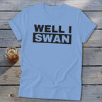 Light blue short-sleeve t-shirt with the text "WELL I SWAN" printed in black on the chest, lying on a wooden surface beside a glass of iced tea.