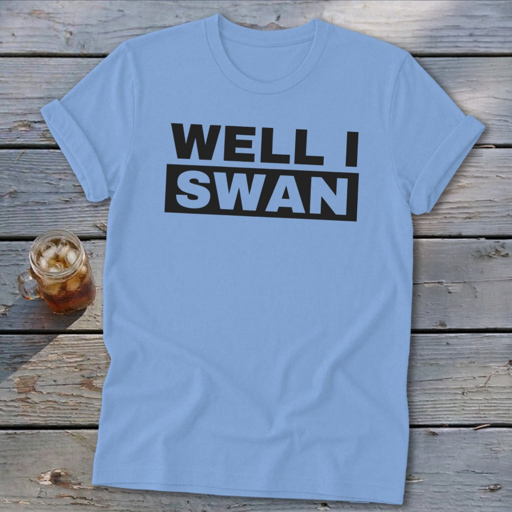 Light blue short-sleeve t-shirt with the text "WELL I SWAN" printed in black on the chest, lying on a wooden surface beside a glass of iced tea.