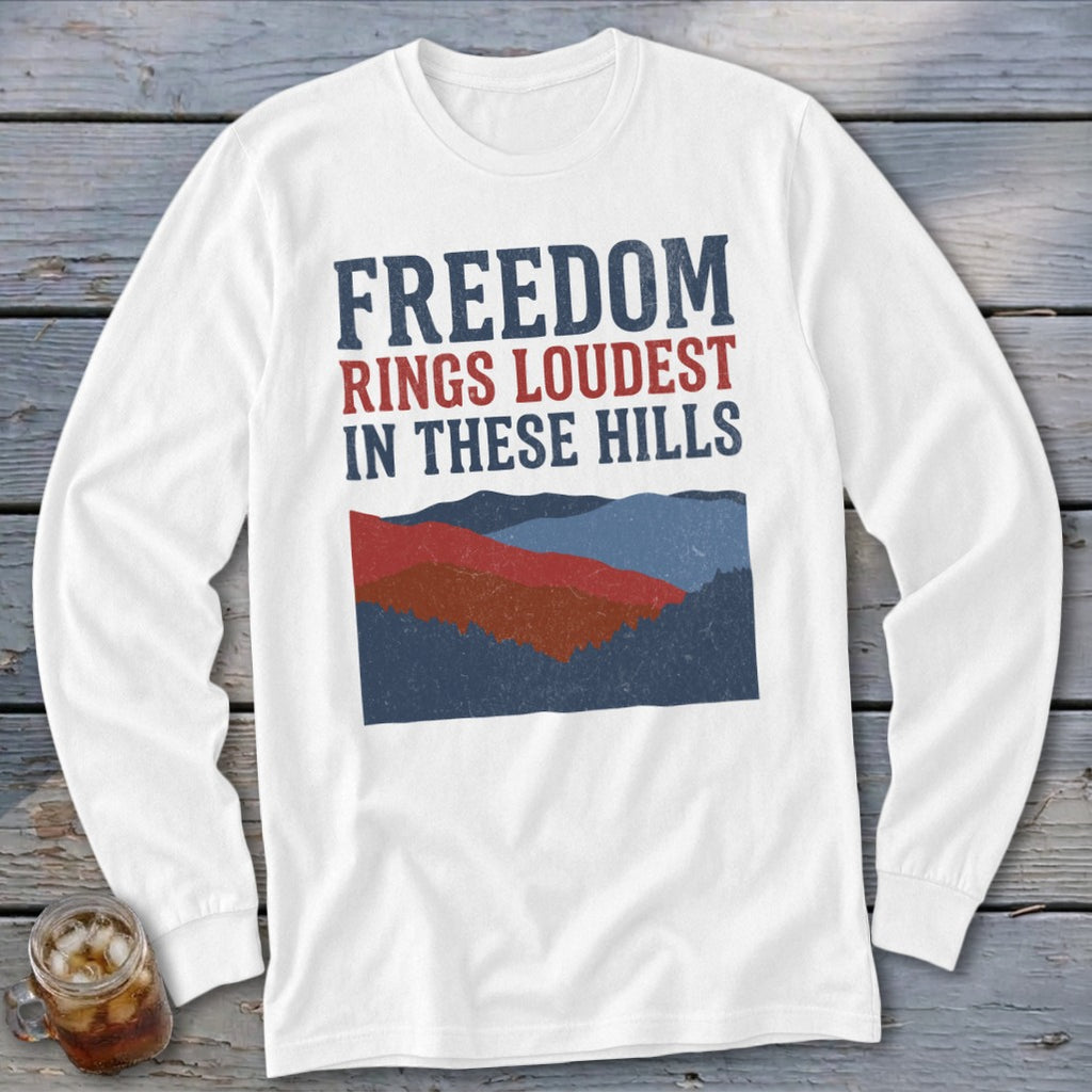 White long-sleeve t-shirt with red and blue graphic text reading "FREEDOM RINGS LOUDEST IN THESE HILLS" above layered mountain silhouettes.